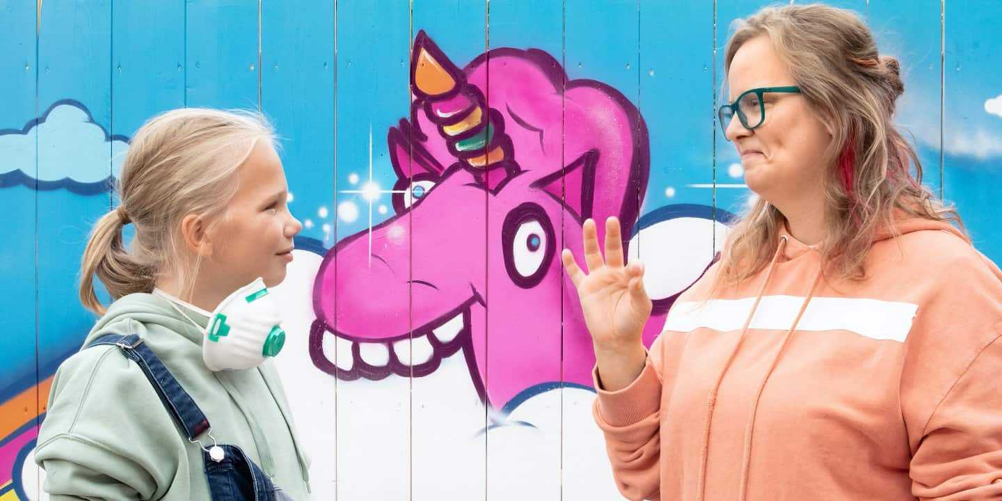 Ein Mädchen und eine gestikulierende Frau blicken sich vor einer bunt bemalten Wand mit einem Einhorn an