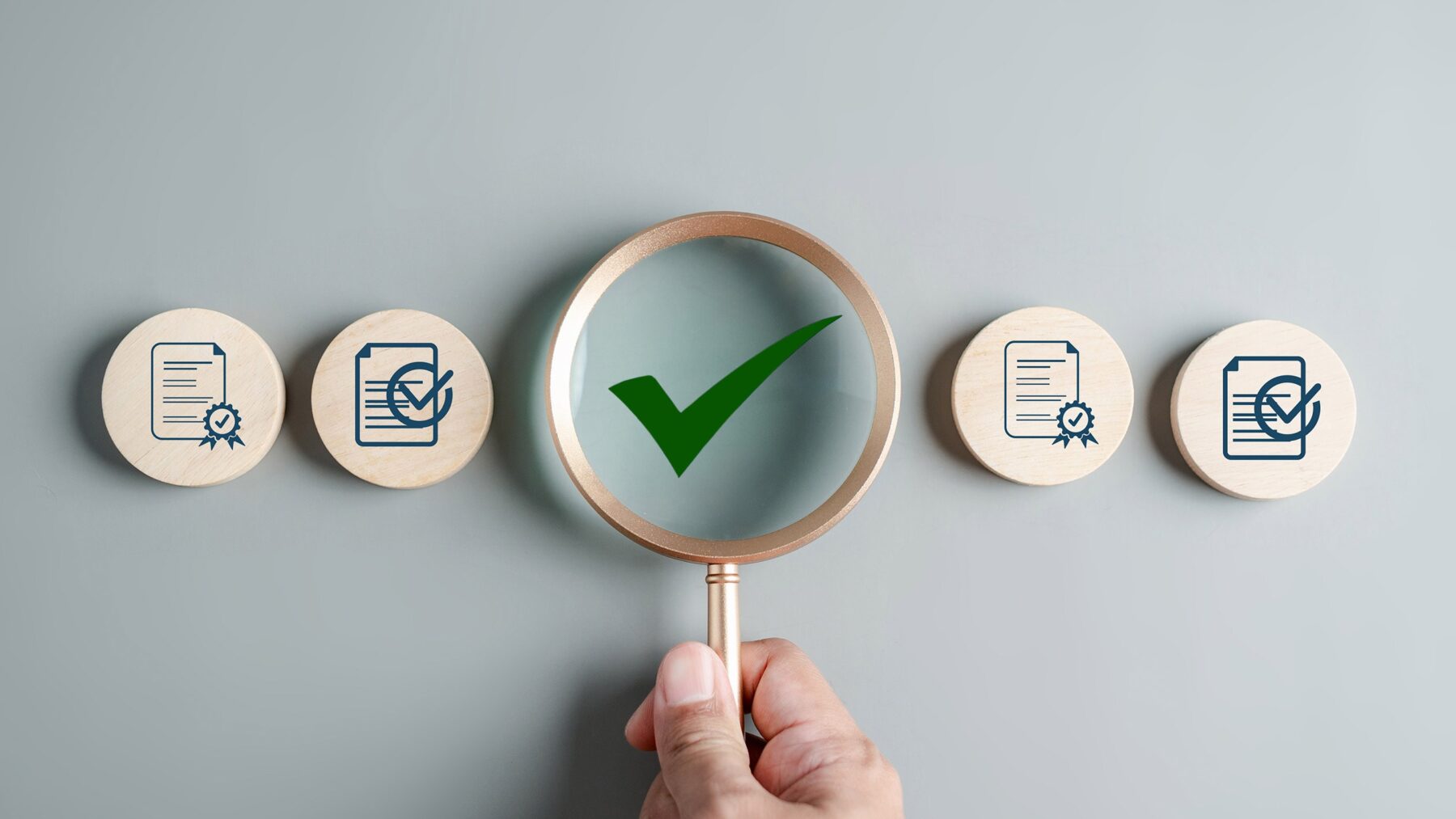 One hand is holding a magnifying glass with a green tick in it. To the left and right of it are small, round wooden plates with a checklist and documents on them.