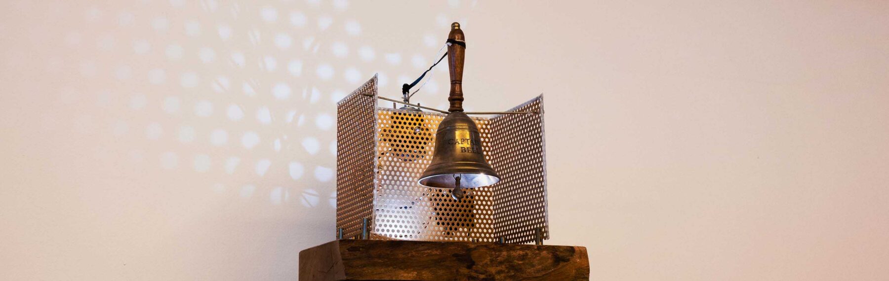 An old sailor's bell made of brass with a wooden handle on a wooden pedestal on which the words "Beer Captain" are engraved. The bell is wired to a motor.