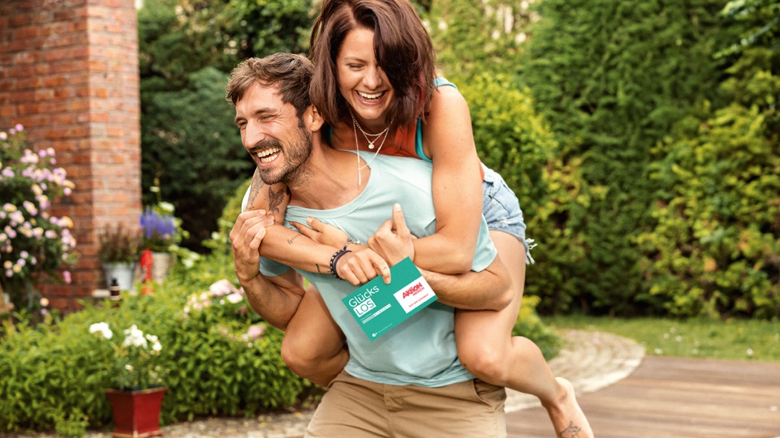 A woman sits piggyback on a man's back in the garden. They are both laughing. She is holding a lucky ticket from Aktion Mensch.