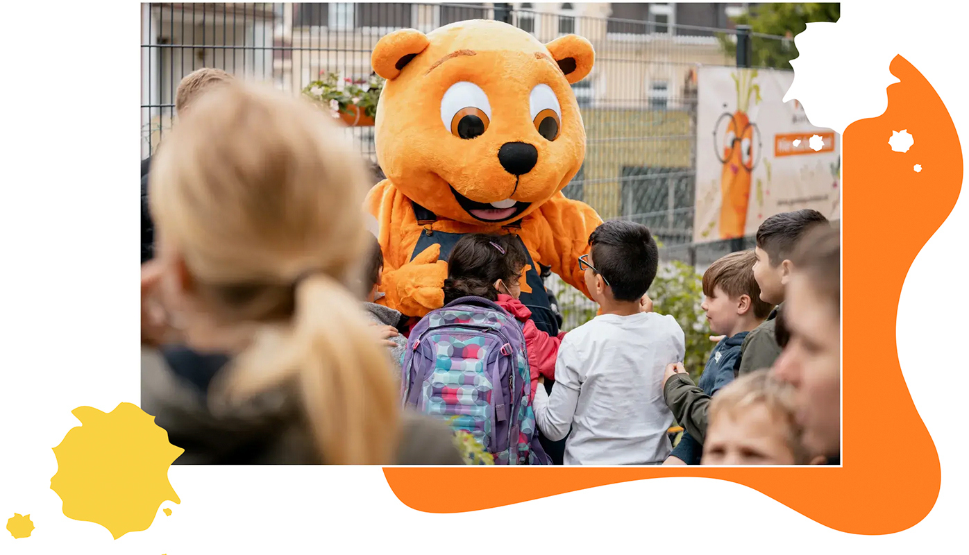 Ein großer OBI-Biber wird auf einem Schulhof von mehreren Schulkindern umringt. Ein Mädchen mit dunklen Haaren umarmt den Biber.