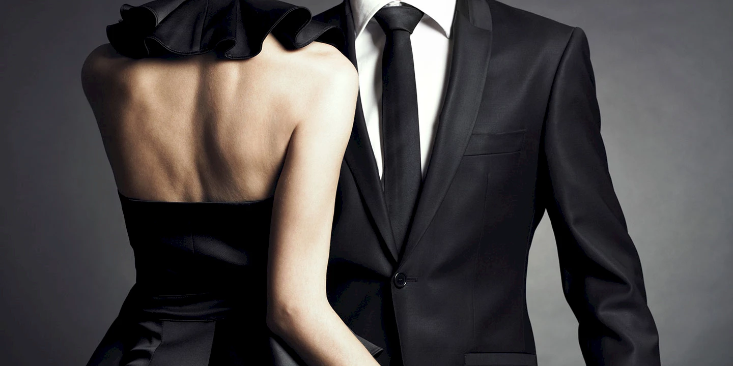 A woman in a black off-the-shoulder evening dress from behind, a man in a black suit with tie from the front. Only the upper parts of their bodies are visible.