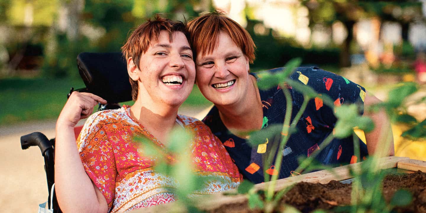 Portrait zweier Frauen in einem grünen Außenbereich. Sie legen ihre Köpfe eng aneinander und lachen in Richtung Kamera.
