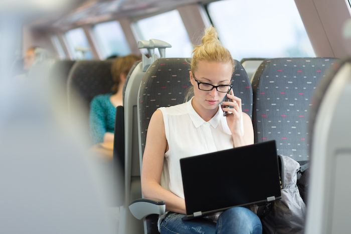 A woman in business attire in a train, holding a phone to her ear and having a laptop on her legs